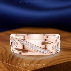 Op de afbeelding: Roségouden ring met een geometrisch ontwerp en een rij kleine, heldere edelstenen. De ring heeft uitgesneden details en is geplaatst op een houten oppervlak met een donkerblauwe achtergrond. De tekst "Solitairetrend" is zichtbaar.