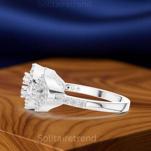 Puede incluir: Un anillo de plata con un grupo de peque&ntilde;as piedras transparentes en un dise&ntilde;o floral. La banda presenta una fila de peque&ntilde;as piedras. El anillo se muestra sobre una superficie de madera con el texto "Solitairetrend".