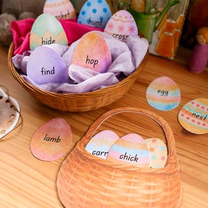 May include: A wicker basket filled with colorful Easter egg cutouts, some with words like "hide" and "hop." Another basket holds more eggs, and individual eggs with words like "lamb" and "egg" are scattered around. A children's book is in the background.