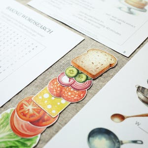 Puede incluir: Ilustraciones de papel de ingredientes para s&aacute;ndwiches, incluyendo pan, lechuga, tomate, queso, salami, pepino y cebolla roja. Tambi&eacute;n hay art&iacute;culos relacionados con la reposter&iacute;a, como un batidor, un colador y una sart&eacute;n, junto con un juego de b&uacute;squeda de palabras.