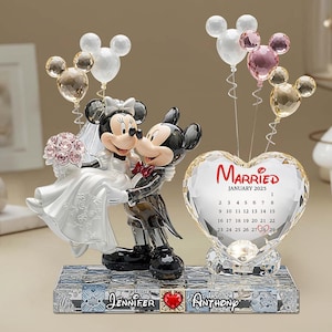 Puede incluir: Un adorno para pastel de bodas con Mickey y Minnie Mouse con atuendo de boda. Minnie sostiene un ramo y Mickey la lleva. Un calendario de cristal en forma de corazón muestra "Casados ​​enero de 2025". También hay globos con cabezas de Mickey Mouse.