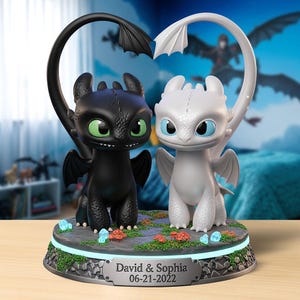 May include: A decorative figurine featuring two dragon figures, one black and one white, with heart-shaped tails. The base includes the names "David & Sophia" and the date "06-21-2022". The dragons have large eyes and are set on a base with faux plants and glowing accents.