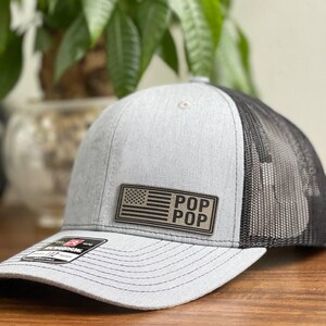 May include: Gray and black trucker hat with a patch featuring an American flag and the words "POP POP". The hat has a curved brim and a mesh back.