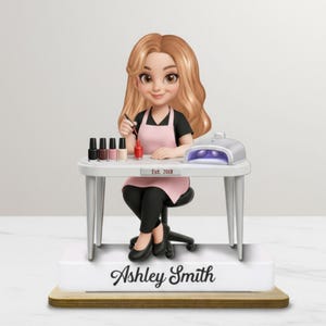 May include: A figurine of a nail technician at a table with nail polish bottles and a UV lamp. The figurine is wearing a pink apron and black clothing. The base of the figurine has the name "Ashley Smith" and "Est. 2018".