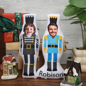 Op de afbeelding: Twee gepersonaliseerde pluche notenkrakerpoppen. De ene pop is gekleed in grijs en zwart, de andere in blauw en goud. Elke pop heeft een gezicht erop gedrukt. Het woord "Robison" staat op de bodem van één pop.