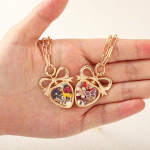 May include: Two gold-toned heart-shaped pendants suspended from matching link chains. Each pendant features a bow-shaped top and a clear centre filled with miniature colourful charms, including a yellow giraffe, a purple butterfly, and flowers.