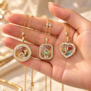 May include: Three gold-tone necklaces featuring clear glass locket pendants framed with small white crystals. The pendants contain various charms including a red heart, an evil eye, and a strawberry. Each chain includes small crystal accents.