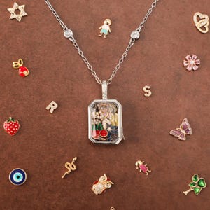 Peut inclure: Un collier médaillon argenté avec un pendentif rectangulaire en verre rempli de petites breloques, dont des cerises, des fleurs et des cristaux. La chaîne comporte deux pierres rondes transparentes. Diverses petites breloques entourent le collier.