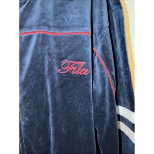 May include: Navy blue velour track jacket with red trim and the word "Fila" embroidered in red script. The jacket has white stripes on the sleeves.