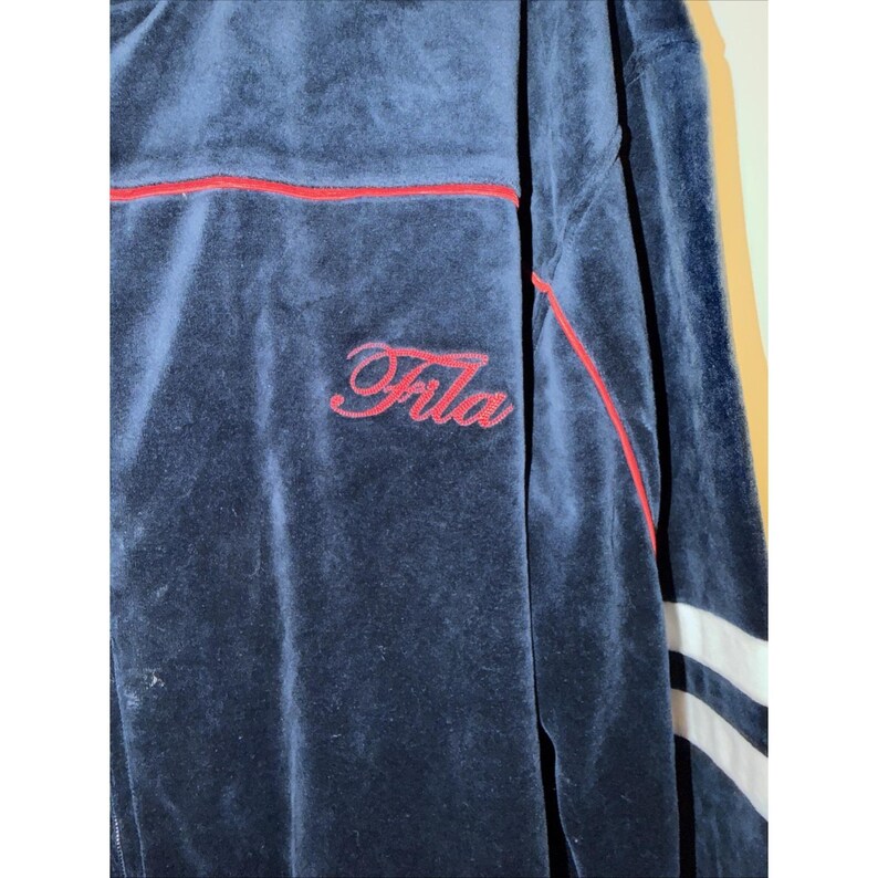 May include: Navy blue velour track jacket with red trim and the word "Fila" embroidered in red script. The jacket has white stripes on the sleeves.
