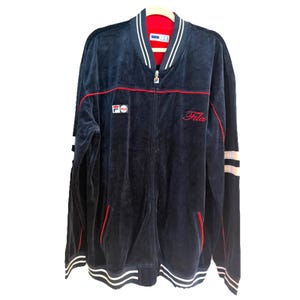 May include: Navy blue zip-up jacket with red and white accents. The jacket features the Fila logo on the chest and the word "Fila" in red script. The jacket has a red interior and white stripes on the sleeves and bottom.