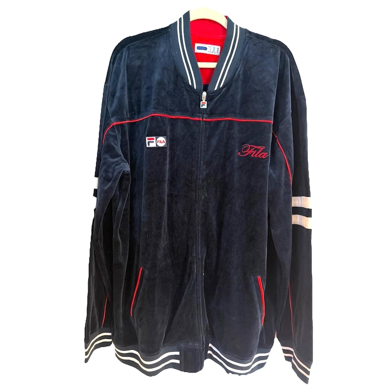May include: Navy blue zip-up jacket with red and white accents. The jacket features the Fila logo on the chest and the word "Fila" in red script. The jacket has a red interior and white stripes on the sleeves and bottom.