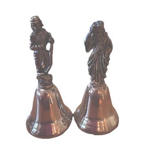 May include: Two antique bronze-coloured bells. Each bell features a detailed figure atop its handle. One figure is a person holding a staff, and the other is a person with a draped head covering. The bells have a polished finish.