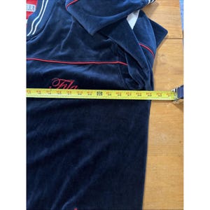 May include: Navy blue velour Fila tracksuit top with red and white accents. The top features the Fila logo in red script. A measuring tape shows the length of the top is approximately 30 inches.