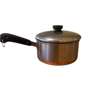 May include: A stainless steel saucepan with a copper base and a black handle. The pan has a lid with a black knob. The handle has a hole for hanging. Kitchenware.