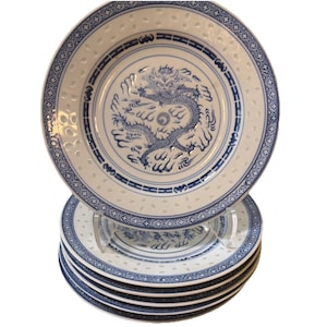 May include: A stack of five white ceramic plates featuring a blue dragon illustration in the centre. The rim of each plate is decorated with a blue geometric pattern and a series of small, embossed circular indentations around the edge.