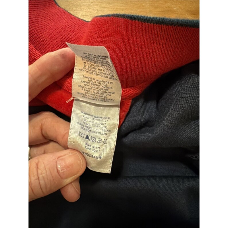 May include: Close-up of a garment tag with care instructions. The tag is white with black text and features washing symbols. The garment has a red collar and navy blue fabric.