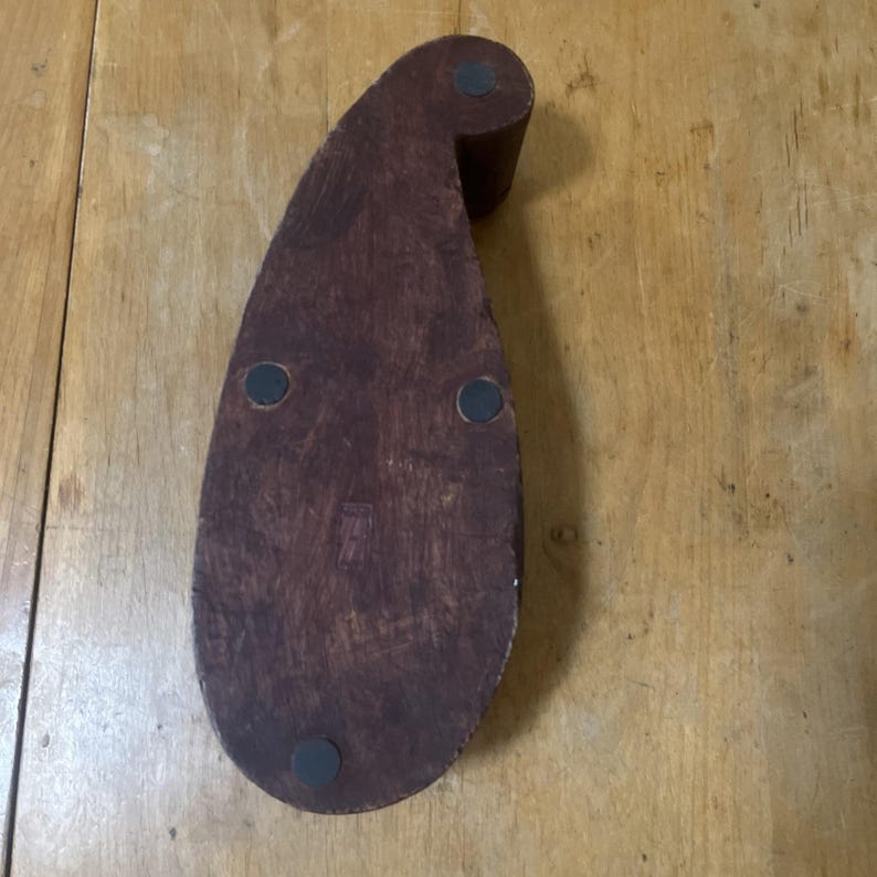 May include: A dark brown wooden object with a teardrop shape and a rounded top. The object has three black circular pads on the bottom and a small cylindrical piece at the top. The wood grain is visible.