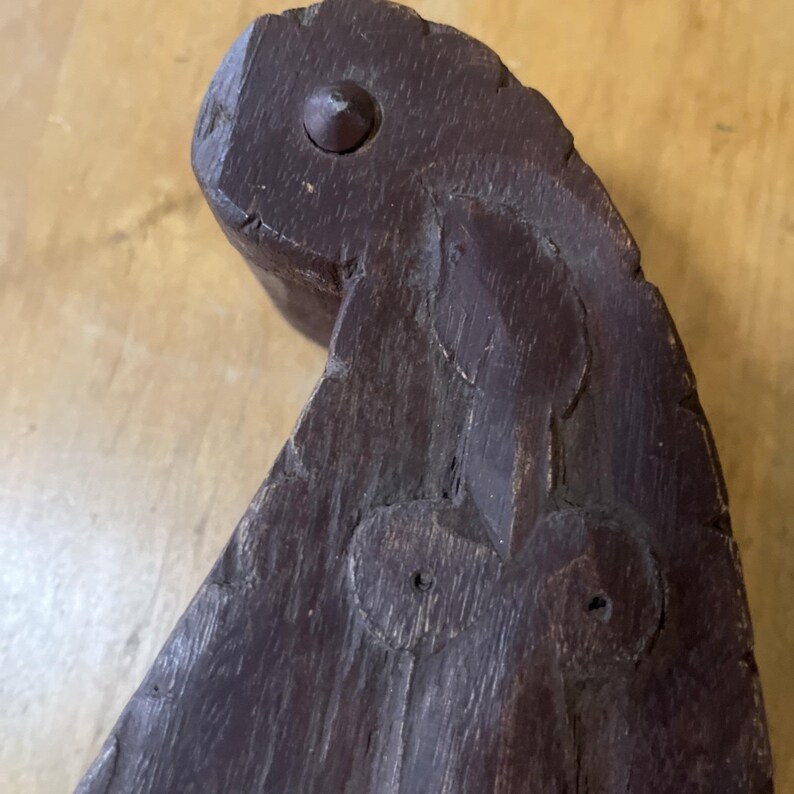 May include: Close-up of a dark brown, carved wooden object with a rounded top and a pointed end. The object has a textured surface and two circular indentations. A small wooden peg is visible at the top. The object appears to be a decorative element.