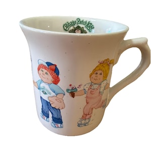 Peut inclure: Tasse en céramique blanche avec des personnages Cabbage Patch Kids. Un personnage porte une casquette de baseball et tient un gant de baseball, l'autre tient un cornet de crème glacée. La tasse a une anse incurvée et le logo Cabbage Patch Kids.