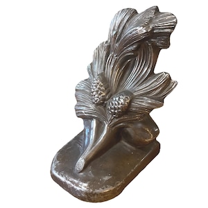 May include: A silver-coloured metal sculpture of a pine branch with two pine cones. The sculpture has a rectangular base and is designed with detailed textures. The overall design is reminiscent of nature.