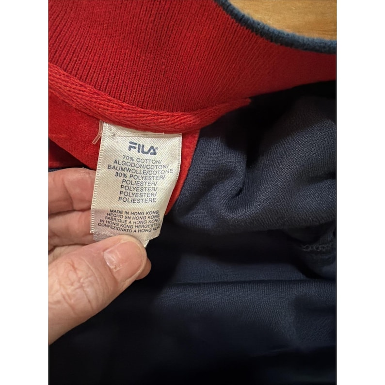 May include: Close-up of a navy blue and red FILA sweatshirt. The red collar and cuffs contrast with the navy body. A white tag with the FILA logo and fabric composition is visible.