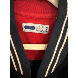 May include: Close-up of a navy blue and red jacket with white stripes on the sleeves. The jacket has a red collar and a tag with the brand name "FILA" and size information.