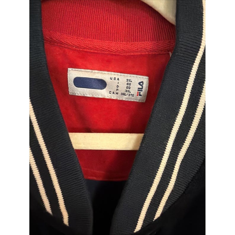 May include: Close-up of a navy blue and red jacket with white stripes on the sleeves. The jacket has a red collar and a tag with the brand name "FILA" and size information.