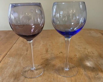May include: Two wine glasses with clear stems and bases. One glass is a deep purple color with white polka dots, and the other is clear with a blue base and white polka dots. The glasses are on a wooden surface.