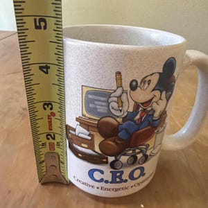 May include: A speckled off-white ceramic mug featuring a graphic of Mickey Mouse sitting at a desk with a computer and phone. The text C.E.O. Creative Energetic Optimistic is printed below. A yellow measuring tape shows the mug is 5 inches tall.