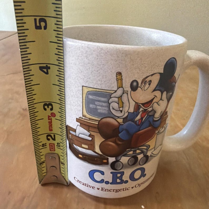 May include: A speckled off-white ceramic mug featuring a graphic of Mickey Mouse sitting at a desk with a computer and phone. The text C.E.O. Creative Energetic Optimistic is printed below. A yellow measuring tape shows the mug is 5 inches tall.