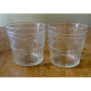 May include: Two clear glass tumblers with a grape vine design. The glasses are cylindrical with a slightly flared rim and a decorative band featuring grape clusters and leaves. The glasses are sitting on a wooden surface.