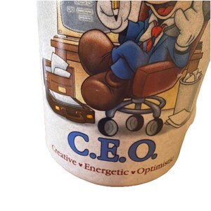 May include: A ceramic mug featuring a cartoon character in a blue suit sitting in an office chair at a desk. The text C.E.O. is printed in blue, with the words Creative, Energetic, and Optimistic printed in smaller brown letters underneath.