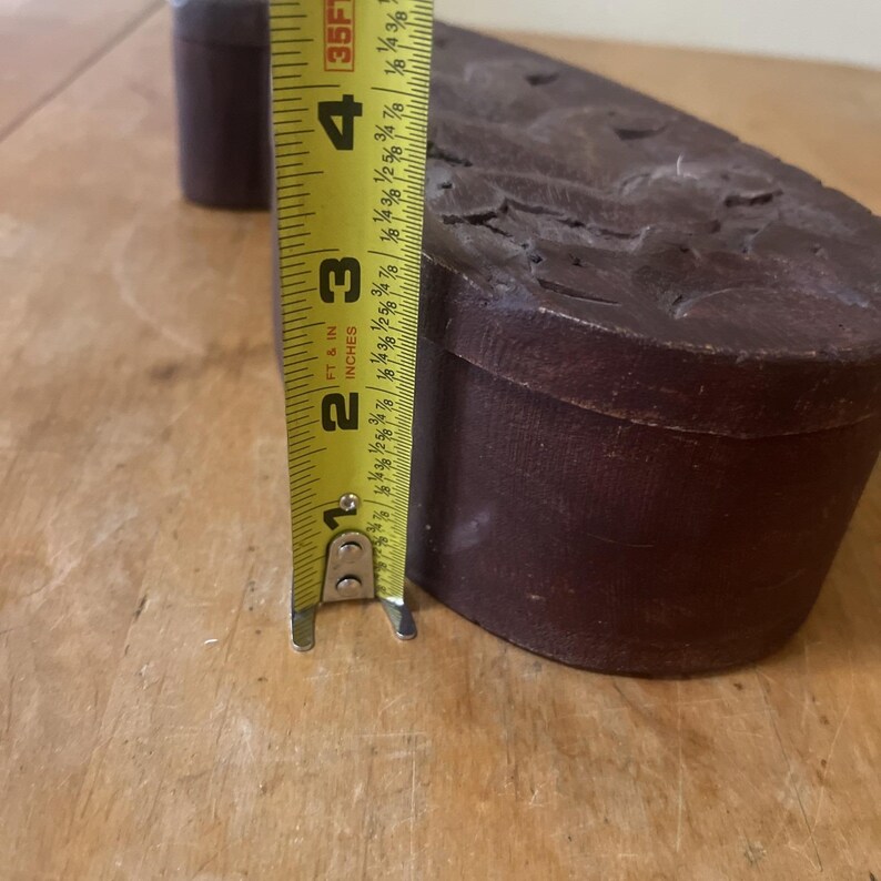May include: A dark brown, rectangular object with a carved surface, possibly a shoe sole or a similar component. The object is measured with a yellow tape measure, showing a height of approximately 3 inches.