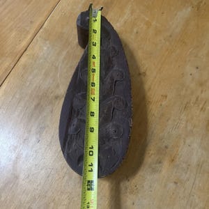 May include: A dark brown, teardrop-shaped wooden carving with a cylindrical handle. The carving features a series of raised, stylized floral or organic shapes. A yellow measuring tape shows the carving is approximately 12 inches long.