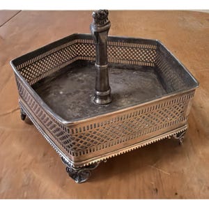 Puede incluir: Una bandeja de servir hexagonal plateada con un diseño de celosía en los lados y pies decorativos. Un asa central con una parte superior en forma de corona se eleva desde el centro de la bandeja. La bandeja está sobre una superficie marrón.