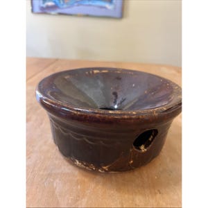 May include: A dark brown ceramic bowl with a wide, flared rim and a small hole on the side. The bowl has a decorative wavy pattern around the base and is sitting on a wooden surface.