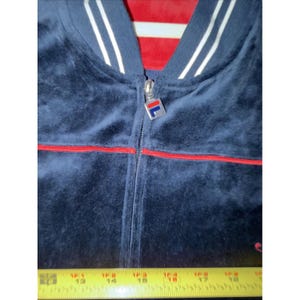 May include: A navy blue velour zip-up jacket with red trim and white stripes on the collar. The jacket features a silver zipper with a logo and a red lining visible at the neckline. The jacket is measured with a yellow tape measure.