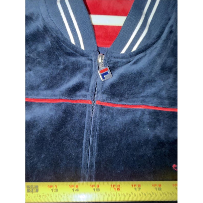 May include: A navy blue velour zip-up jacket with red trim and white stripes on the collar. The jacket features a silver zipper with a logo and a red lining visible at the neckline. The jacket is measured with a yellow tape measure.