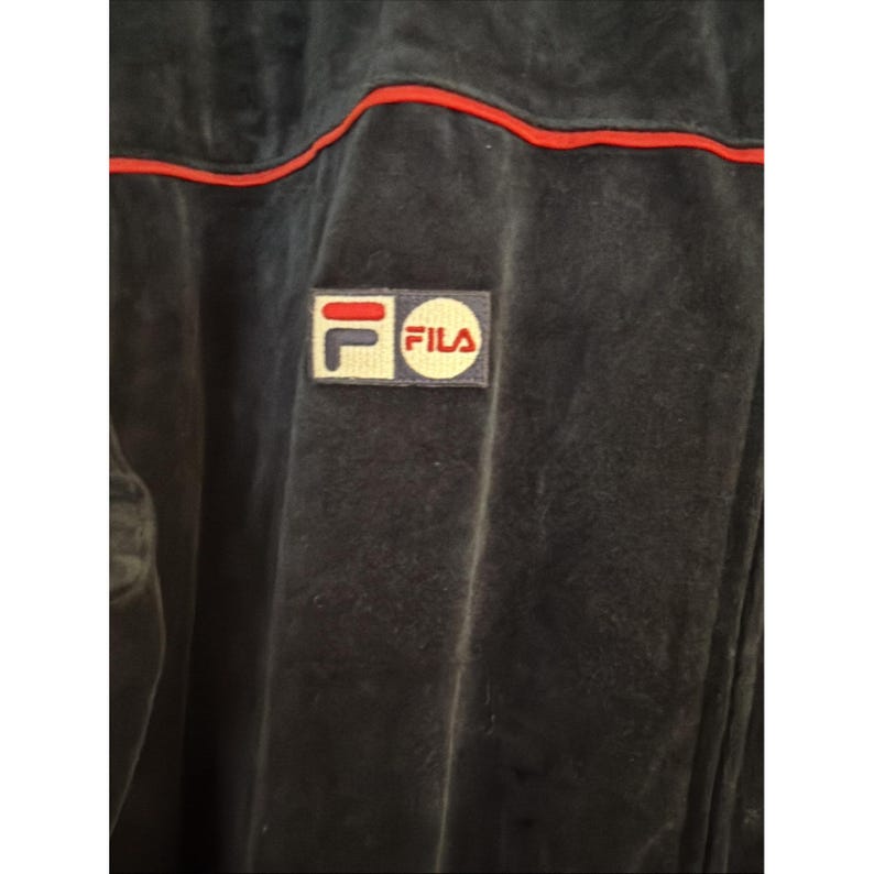 May include: Close-up of a dark navy blue jacket with a red stripe along the seams. The jacket features a small rectangular patch with the FILA logo in red, white, and navy blue. The fabric appears to be a soft, velvety material.