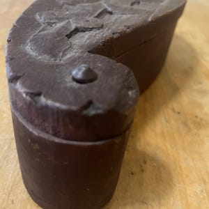 May include: A dark brown, wooden box with a curved, carved lid. The box has a rounded shape and a small, decorative knob on the front. The wood appears aged and has a textured surface.
