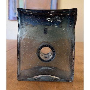 May include: A rectangular, dark blue glass vase with a circular opening. The vase has rounded corners and a textured surface with small bubbles. The vase is sitting on a wooden surface.