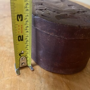 May include: A dark brown, cylindrical object with carved details on the top surface. A yellow tape measure shows the object is approximately 2 inches (5 cm) tall. The tape measure reads "FT & IN INCHES".