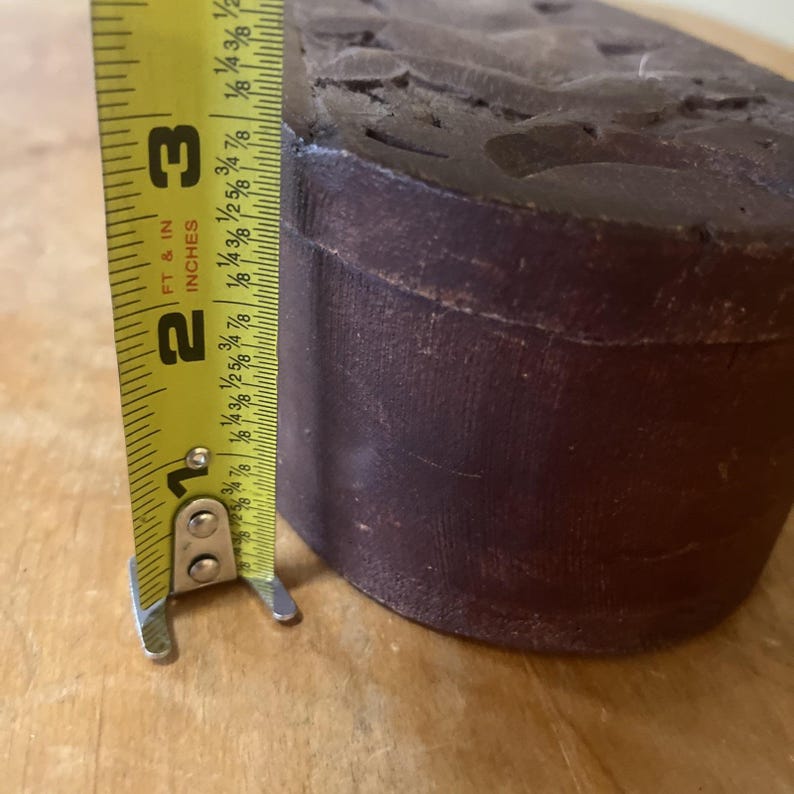 May include: A dark brown, cylindrical object with carved details on the top surface. A yellow tape measure shows the object is approximately 2 inches (5 cm) tall. The tape measure reads "FT & IN INCHES".