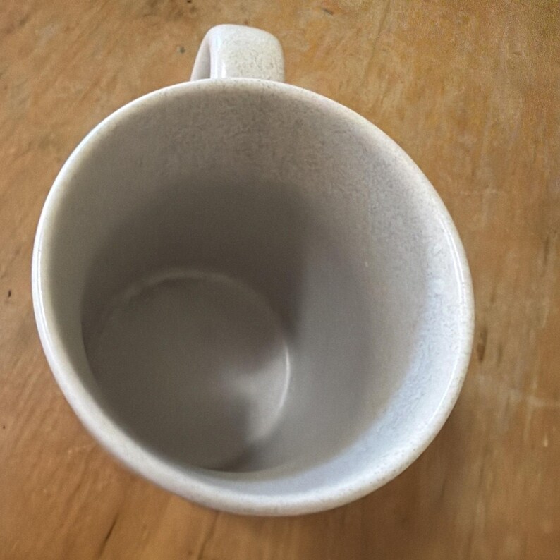 May include: A ceramic mug with a speckled light gray finish viewed from directly above. The mug features a smooth interior surface and a single handle extending from the side. The speckled pattern covers the entire exterior and the rim of the mug.