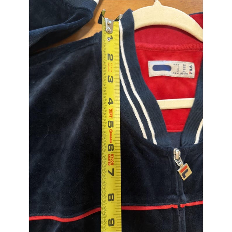 May include: A navy blue velour track jacket with red trim and white stripes. The jacket has a zipper closure and a gold FILA logo. The jacket measures approximately 9 inches in length.