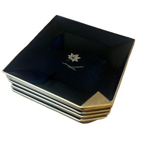 May include: A stack of square, black plates with gold-colored corners. Each plate features a silver leaf and stem design in the center. The plates have a glossy finish and are stacked neatly.