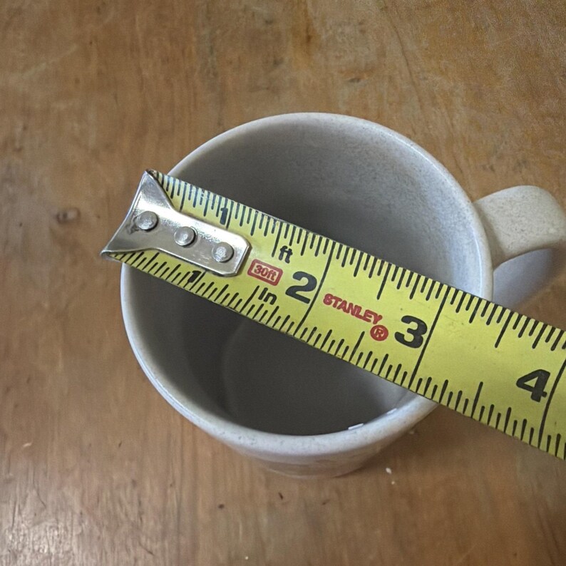 May include: A light beige speckled ceramic mug with a handle, shown from a top-down perspective. A yellow Stanley measuring tape is placed across the opening, indicating a diameter of approximately 3.25 inches for the mug's rim.