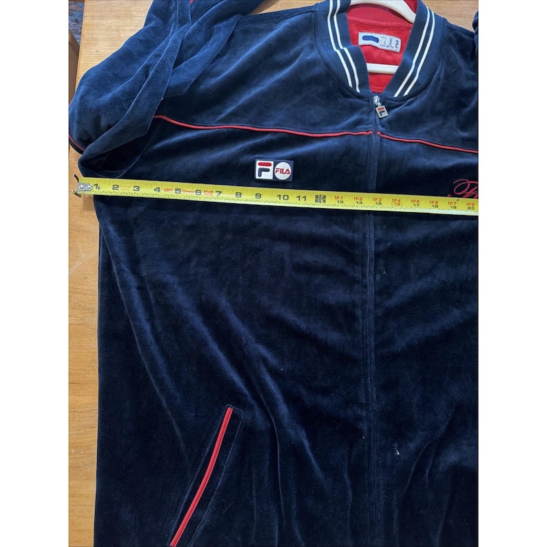 May include: Navy blue velour track jacket with red and white accents. Features a full zipper, red piping, and the Fila logo on the chest. The jacket measures approximately 11 inches across the chest.
