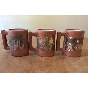 Op de afbeelding: Drie terracotta-kleurige keramische mokken met vierkante handvatten. Elke mok heeft een uniek handgeschilderd ontwerp in bruin-, groen- en goudtinten. De ontwerpen omvatten abstracte patronen en gestileerde afbeeldingen. De mokken hebben een witte binnenkant.